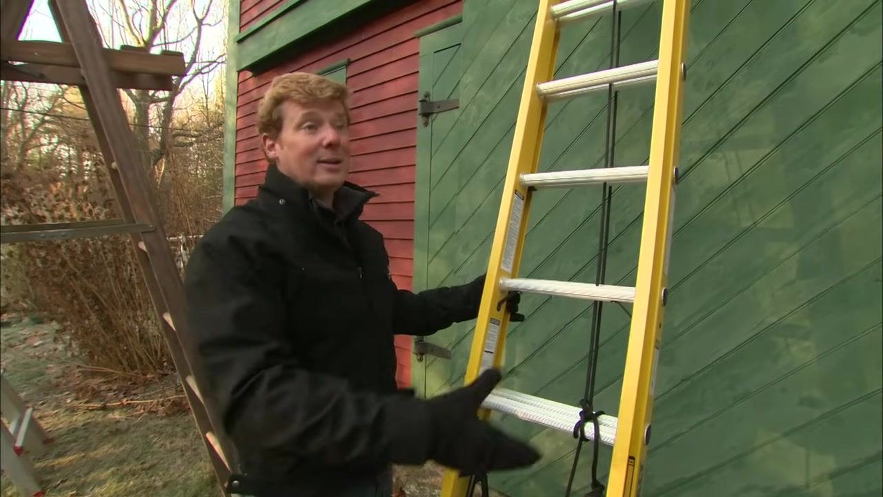 A person in a black jacket points to the rating label on a yellow fiberglass ladder leaning against a green wooden wall.