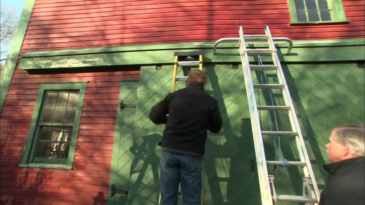 Two people in black jackets inspect an aluminum extension ladder against a red and green barn, checking the rails for bends or creases.