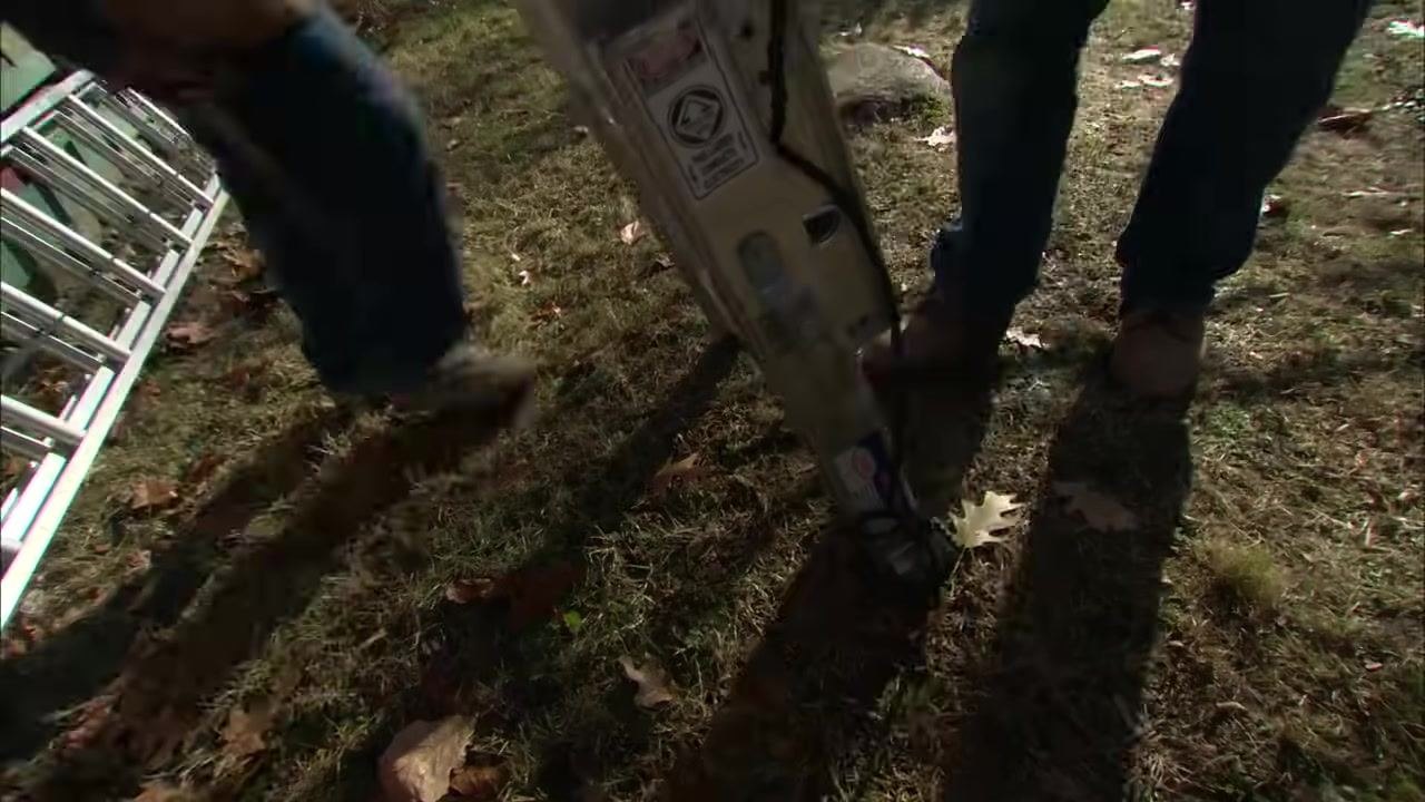 Two people stand near the base of an aluminum extension ladder on grass, checking the safety labels and locking mechanisms.