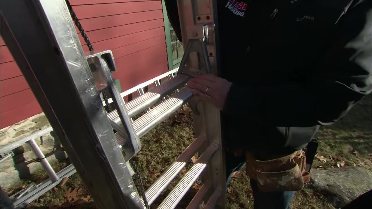 A close-up of a hand engaging the rung locks on an upright aluminum extension ladder, with a red building visible in the background.
