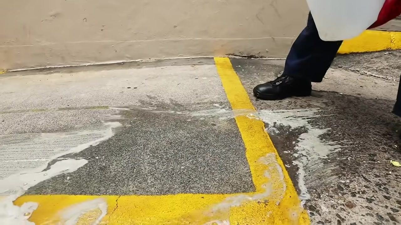 Person in PPE pouring liquid degreaser from a white container onto the cleaned area, preparing for final washdown.