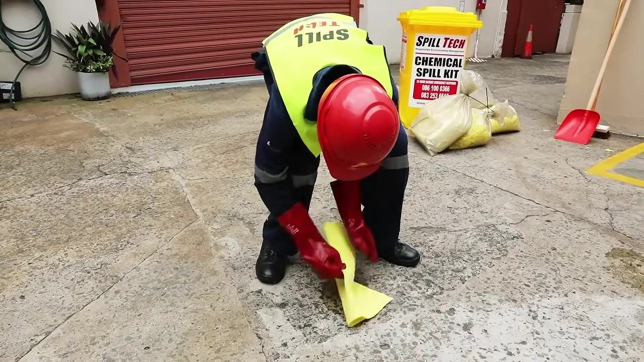Person in full PPE crouched, using a scoop to apply acid neutralizer from a labeled container to a spill surrounded by an absorbent boom, with a container labeled "ACID NEUTRALISER" visible.