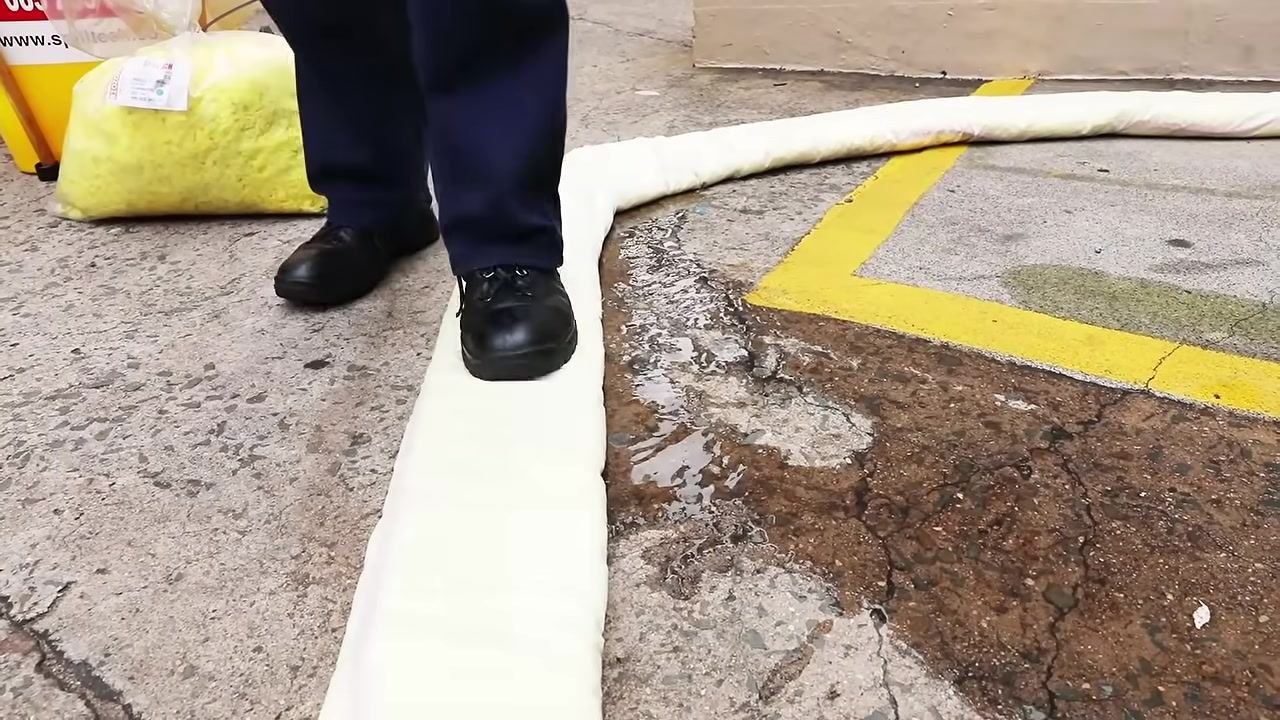 Close-up of feet pressing down on a white absorbent boom placed in a semicircle on concrete to contain a chemical spill, with yellow absorbent material and the kit visible in the background.