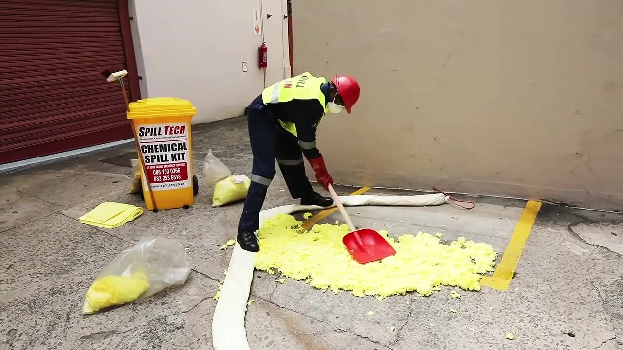Person in full PPE opening a bag of yellow chemical scatter, preparing to apply it to the spill area surrounded by an absorbent boom.