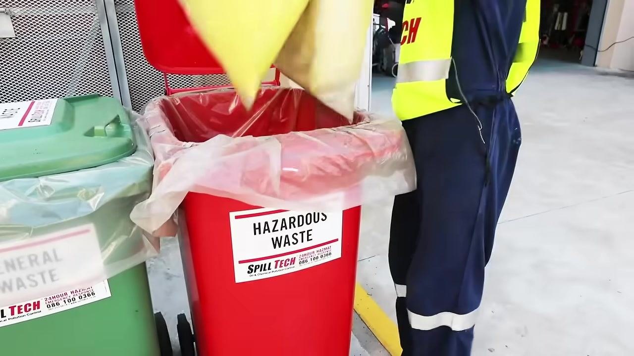 Person in full PPE using a sparkproof spade and broom to sweep up yellow chemical absorbent residue, with the Spill Tech Chemical Spill Kit bin, absorbent pads, and bags visible in an outdoor industrial setting.
