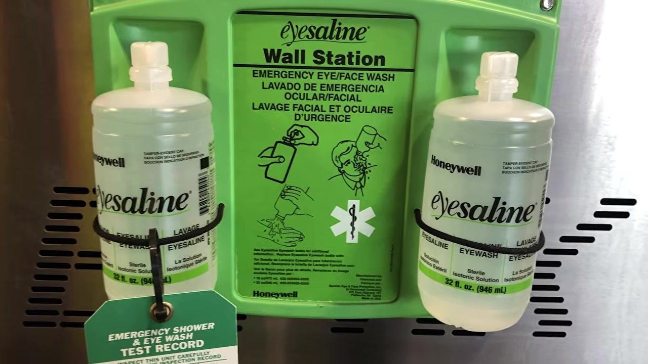 A woman stands next to a green and yellow wall-mounted emergency eyewash station, summarizing the importance of eye safety and injury prevention.
