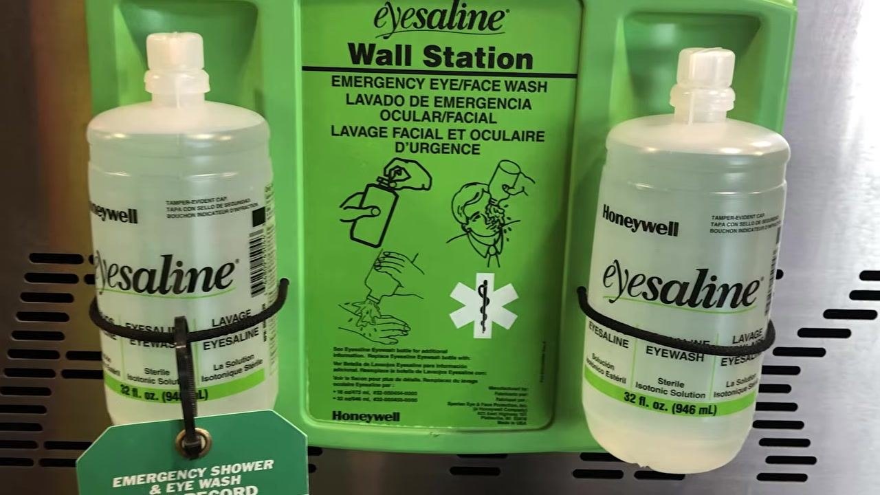 Green emergency eyewash station mounted on a white wall in an industrial facility, with yellow safety railings and clear signage above the station