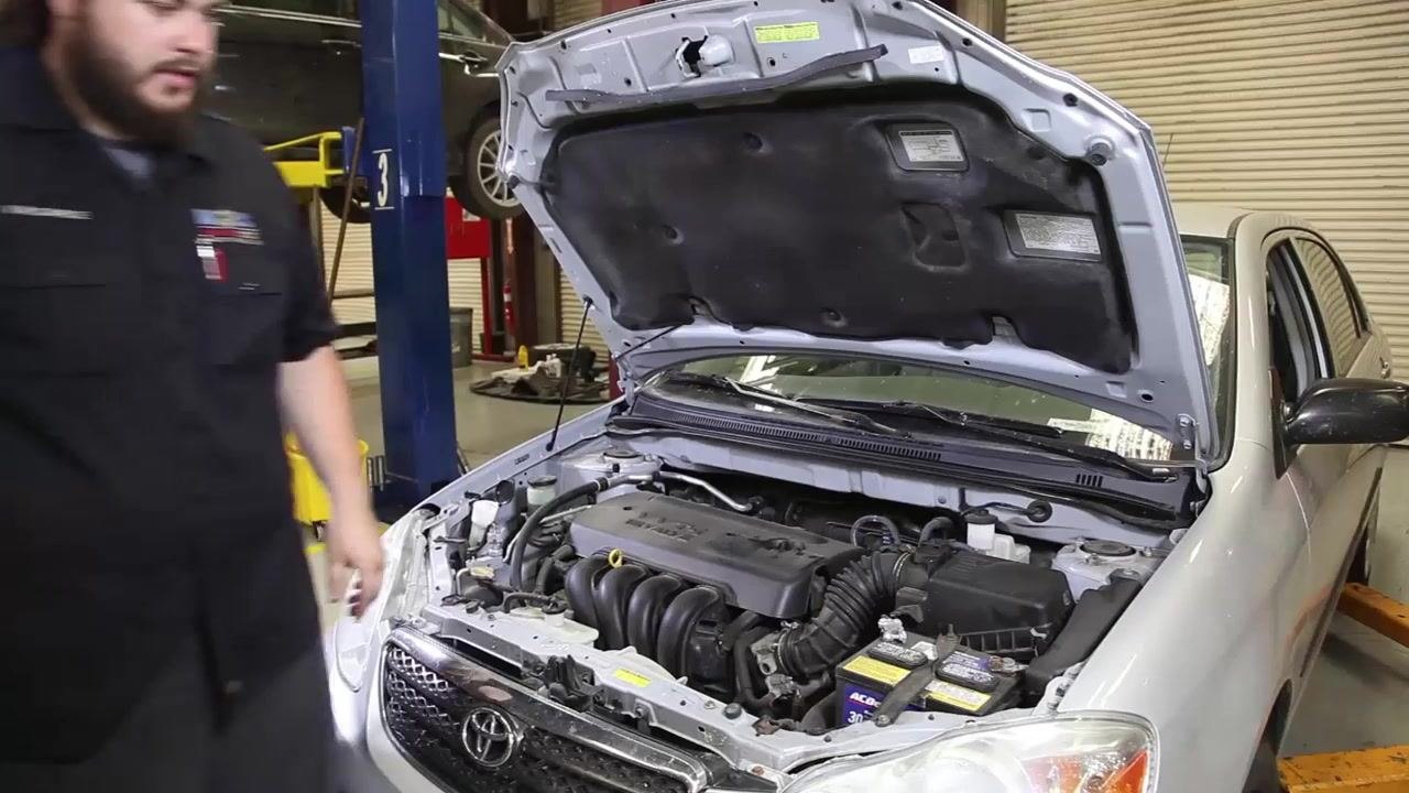 A technician stands at a workbench with a tire pressure gauge, flashlight, power steering fluid, DOT 3 brake fluid, antifreeze coolant, and windshield washer fluid. The technician is in a garage with vehicle lifts and a car with its hood open in the background.