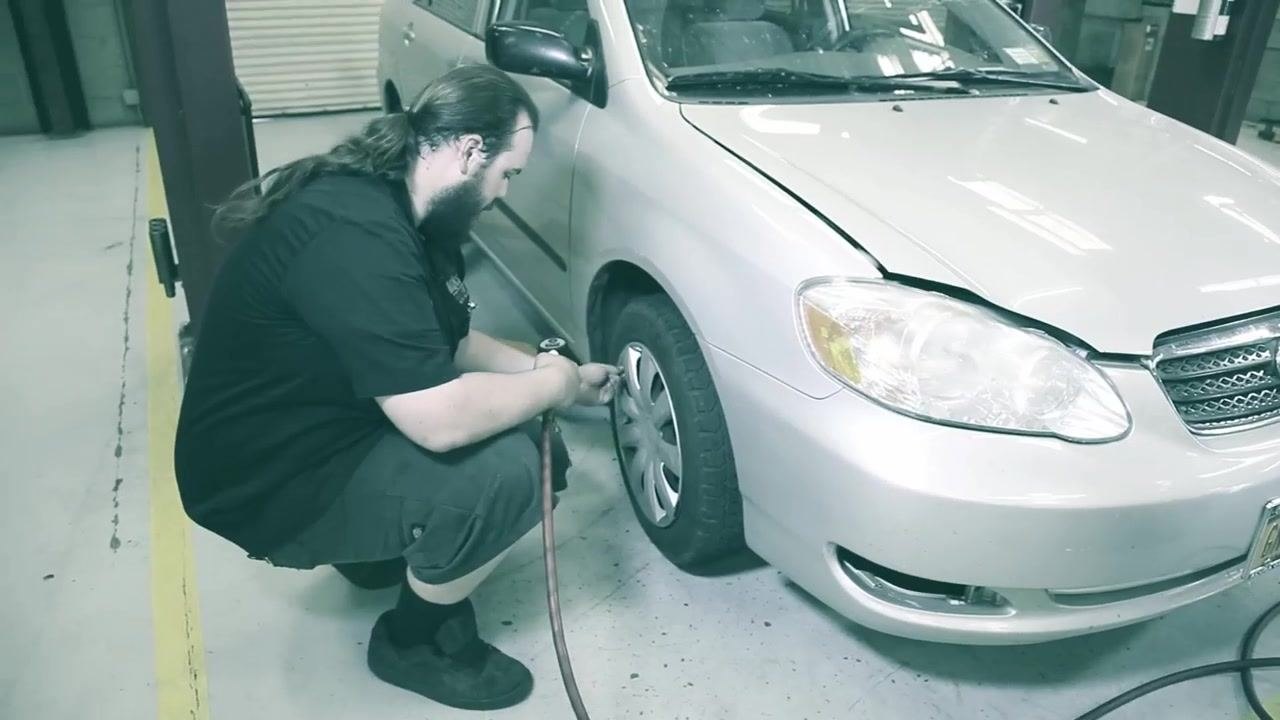 A technician kneels beside the front left tire of a silver car, using a digital tire inflator to check and adjust the tire pressure. The car is in a garage with the hood open.
