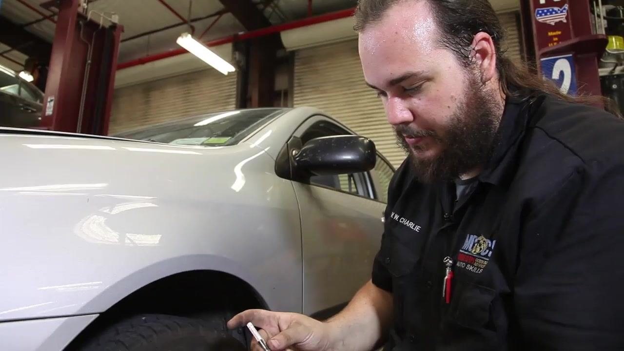A technician kneels beside a car tire in a garage, holding a tire tread depth gauge and preparing to check the tread depth. The technician is focused on the task, and the vehicle is parked inside a well-lit workshop.