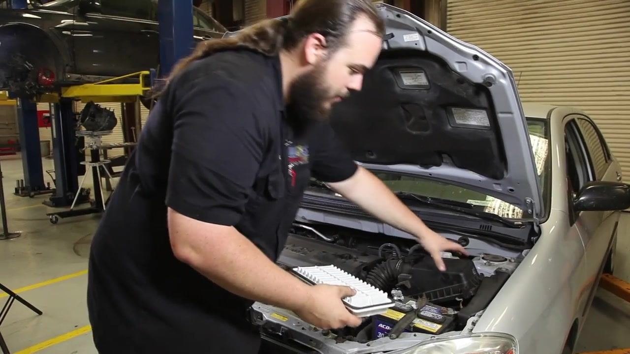 Close-up of a technician pointing at the battery terminals of a Toyota Corolla, highlighting the area to check for corrosion. The battery and engine bay are clearly visible.