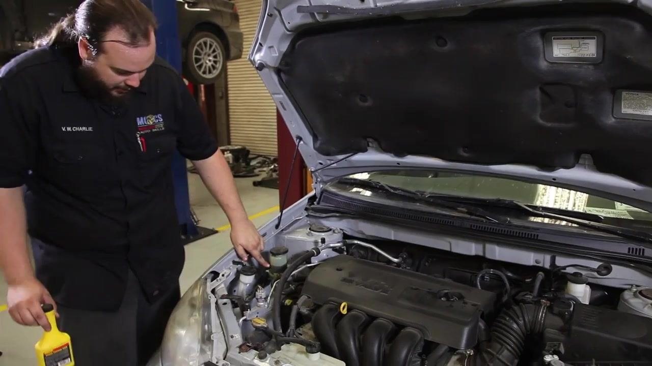 Technician points to the power steering fluid reservoir under the hood of a Toyota Corolla, preparing to open it for inspection. The technician holds a bottle of power steering fluid.
