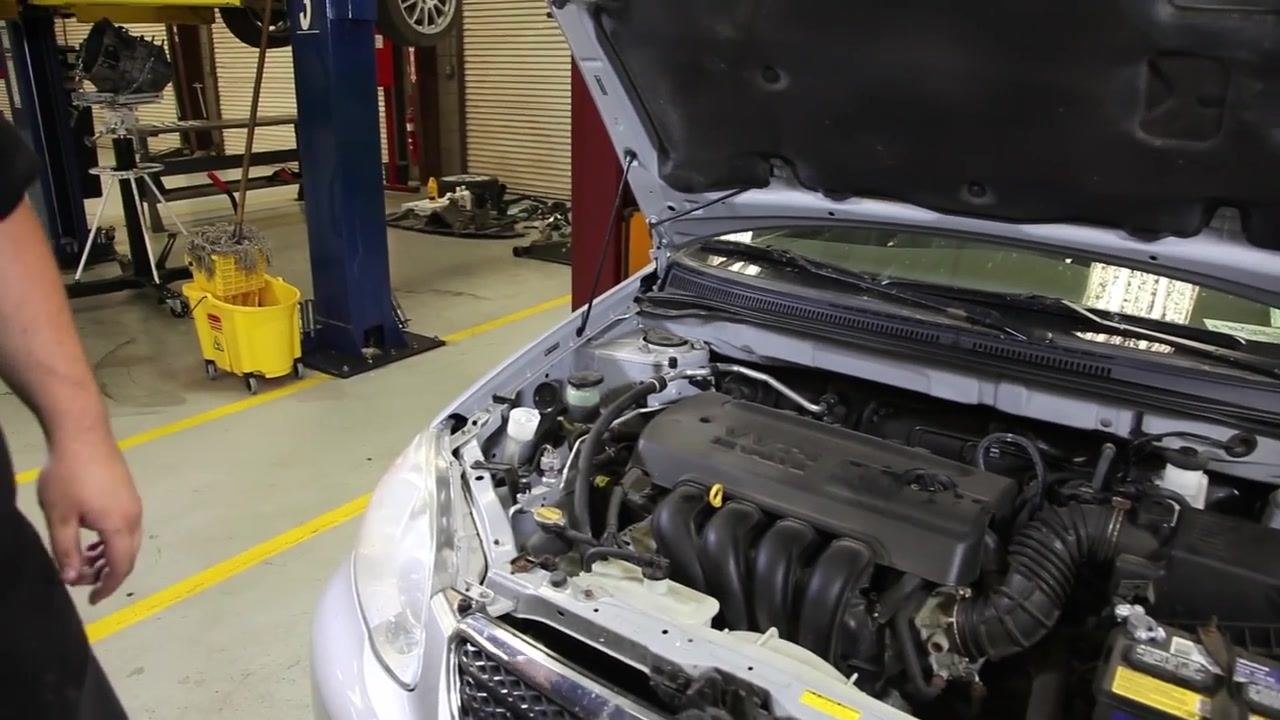 A hand pours power steering fluid into the reservoir under the hood of a car. The fluid is yellowish, and the reservoir is semi-transparent with visible fluid levels. The engine bay and surrounding hoses are visible.