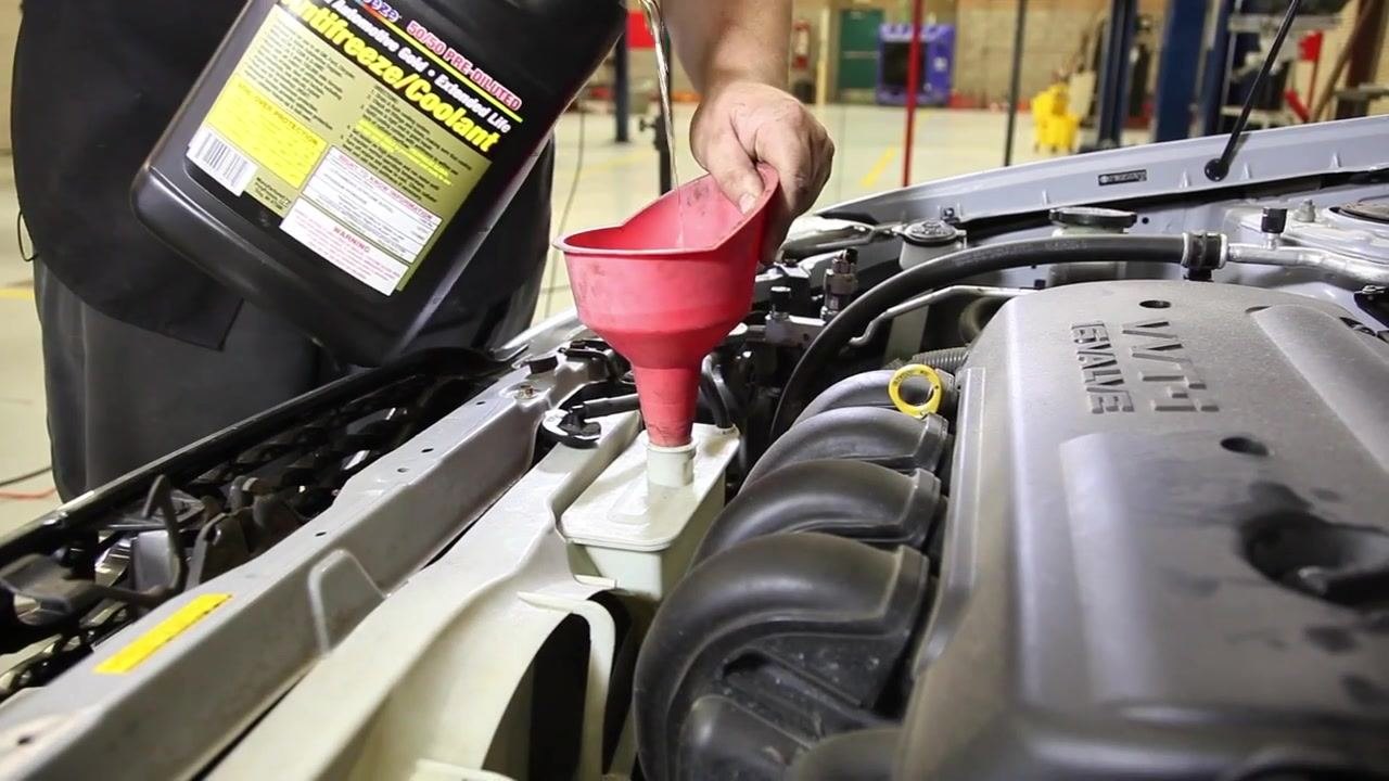 A person holds a jug of blue windshield washer fluid, preparing to pour it into the open reservoir under the hood of a car. The engine bay and other fluid reservoirs are visible.