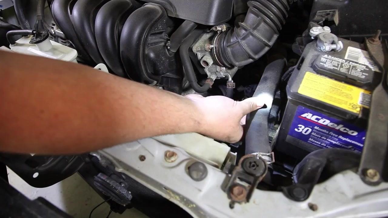 A technician uses a flashlight to inspect and squeeze a radiator hose under the hood of a car. The battery and air intake components are visible nearby.