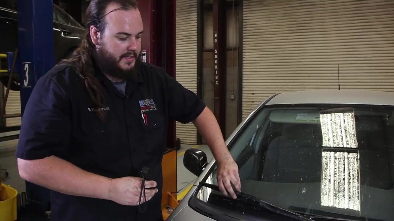 A technician inspects the windshield wiper blade on a car, checking for cracks or tears. The technician is standing in a garage next to the vehicle, with one hand on the wiper and the other pointing at it.