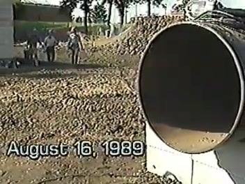 Outdoor construction site showing a large pipe and a job date of August 16, 1989, with three workers visible in the background.