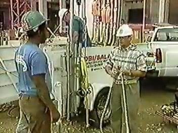 Supervisor in a white hard hat demonstrating rope handling to a group of workers, with safety equipment visible in the background.
