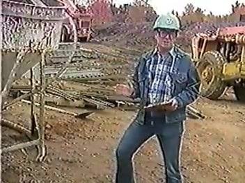Worker in a hard hat and glasses holding a clipboard at a construction site, surrounded by construction equipment and materials.