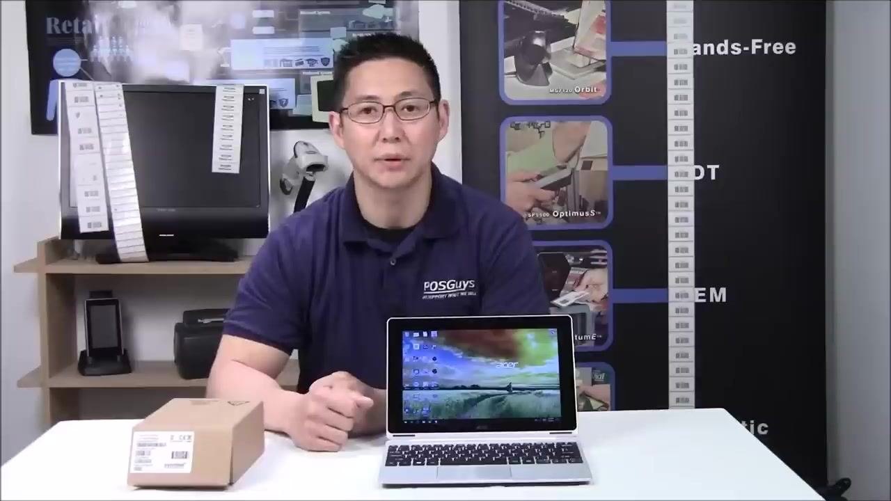 A person sitting at a table with a boxed barcode scanner, a laptop, and various barcode-related equipment in a retail or office setting.