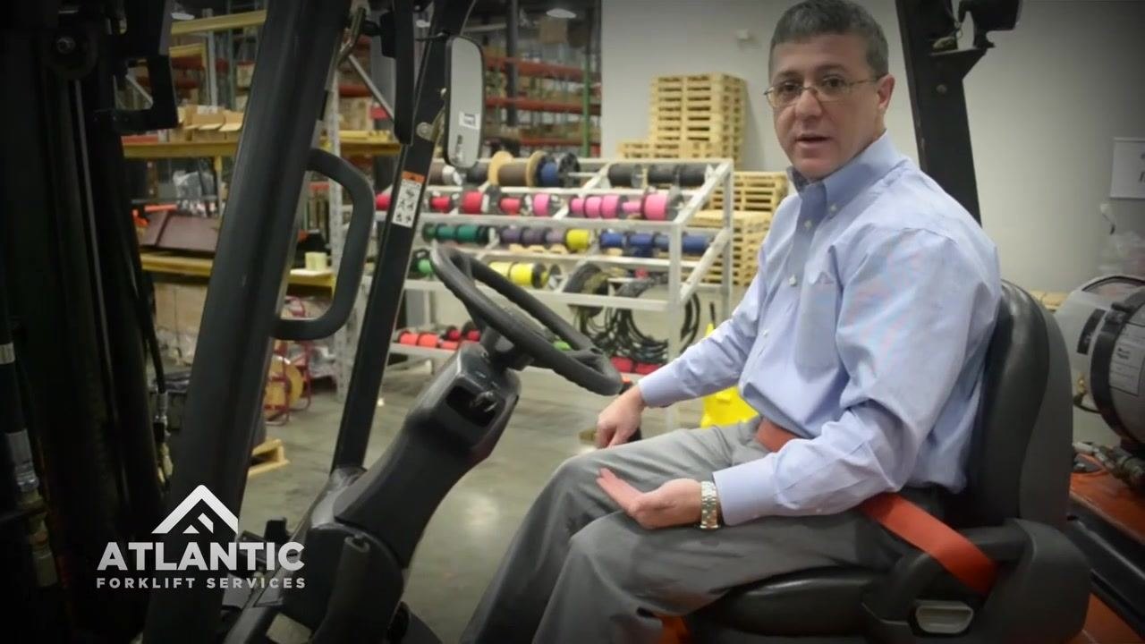 Operator seated in the forklift, demonstrating hydraulic control operation, with visible steering wheel, levers, and seat belt fastened.