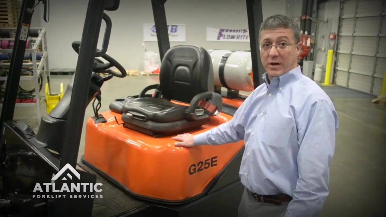 Inspector checking the LP tank on the back of an orange DOOSAN forklift, verifying secure mounting and condition.