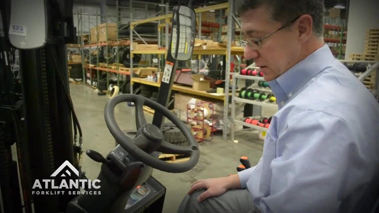 Operator seated in the forklift, hand on controls, visually confirming the parking brake holds with the lift secure in a warehouse setting with shelves and equipment in the background.