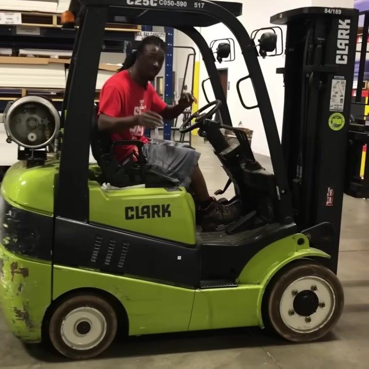 Operator in forklift, speaking directly to camera, warehouse shelving and control panel visible.