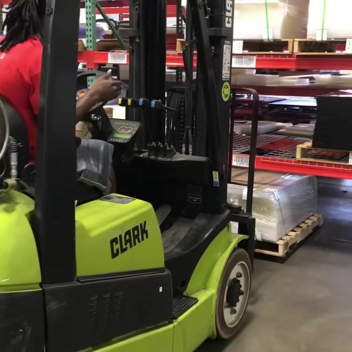 Operator seated in a green CLARK forklift, hand on hydraulic control levers, lowering a pallet inside a warehouse with red shelving and various materials stored on pallets.