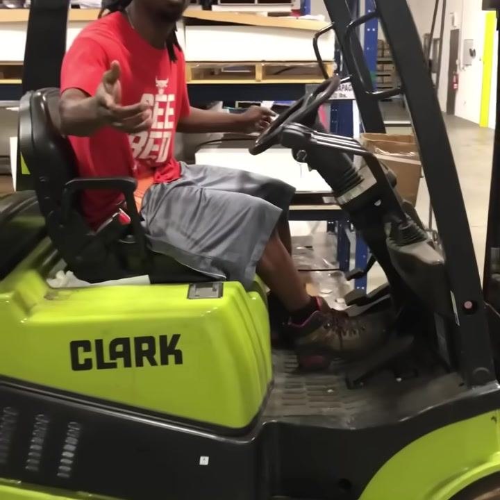 Operator seated on a green CLARK forklift, gesturing towards the control levers. Warehouse shelves and equipment visible in the background.