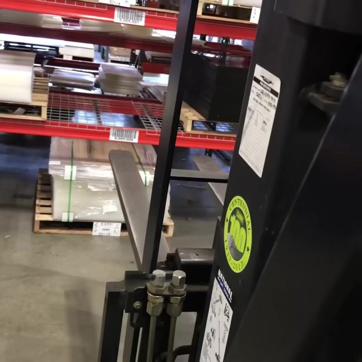 Forks of the forklift visibly shifted to the side, demonstrating the side shift lever's effect. Warehouse shelving and pallets in the background.