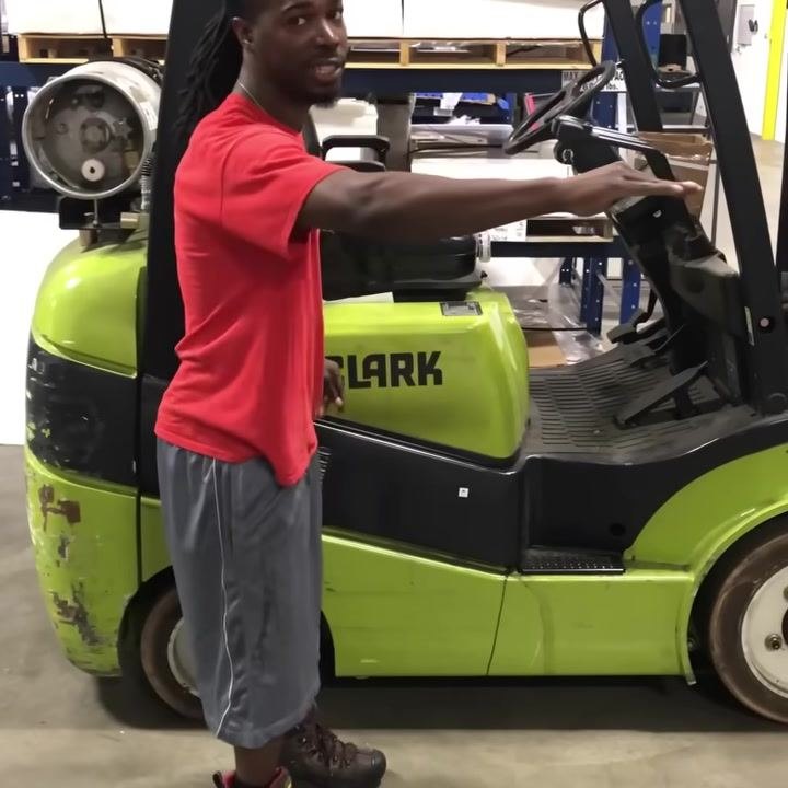 Operator standing next to a green CLARK forklift, pointing at the lever controls while explaining their functions. Warehouse shelving and equipment visible in the background.