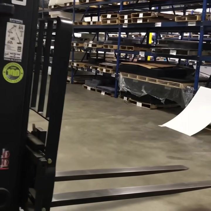 Forks in raised position, mast and safety labels visible, warehouse shelving in background.