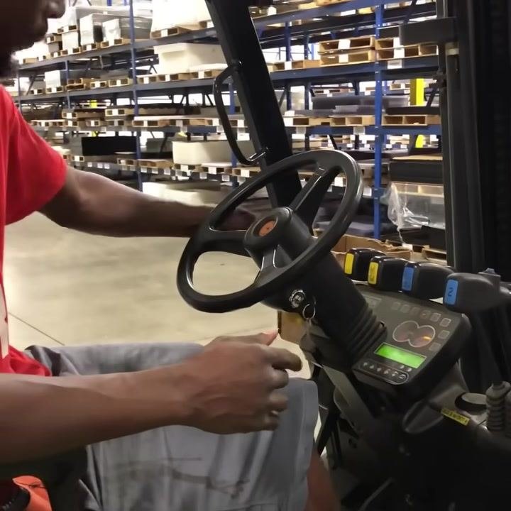 Operator seated in forklift, hand on steering wheel, dashboard and hydraulic levers visible, warehouse shelving in background.
