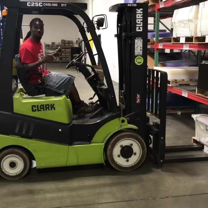 Operator seated in forklift, hands on controls, forks positioned under pallet, warehouse shelving visible.