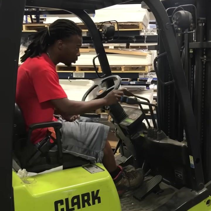 Operator seated in forklift, hand on third hydraulic lever, dashboard and control panel visible, warehouse shelving in background.