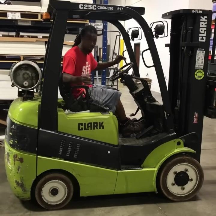 Forklift carrying a large pallet, operator driving straight toward warehouse racking, open warehouse floor visible.