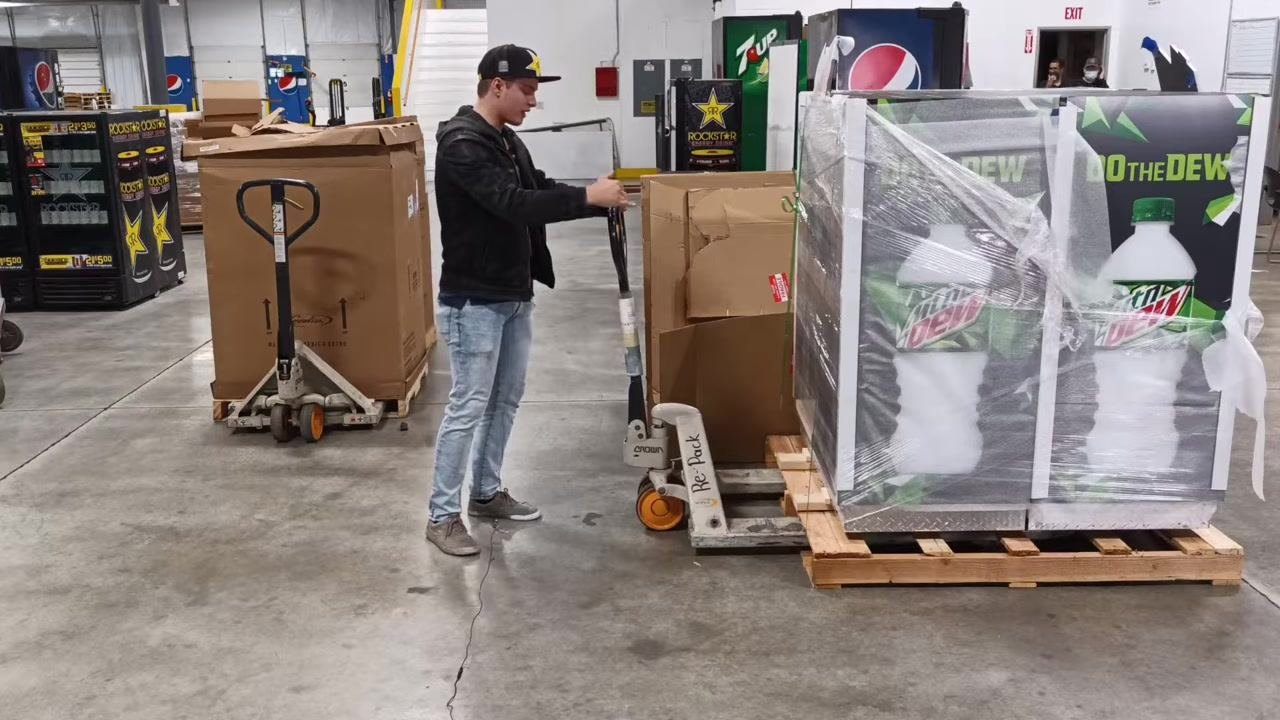 A person positions the pallet jack forks into a pallet, preparing to lift. The warehouse setting includes other pallets and vending machines.