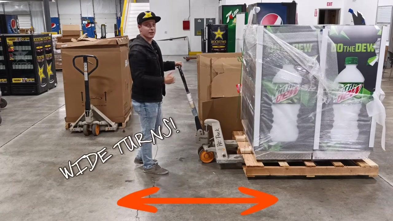 A person stands with the pallet jack handle, ready to crank and lift the pallet. The forks are fully inserted under the pallet.
