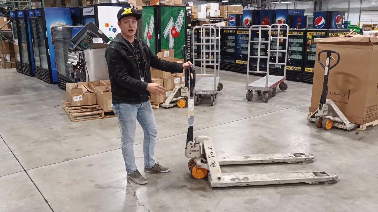 A person demonstrates the danger of the front wheel of a pallet jack in a warehouse. The individual gestures toward the wheel, emphasizing its solid construction. Boxes, vending machines, and other warehouse equipment are visible.