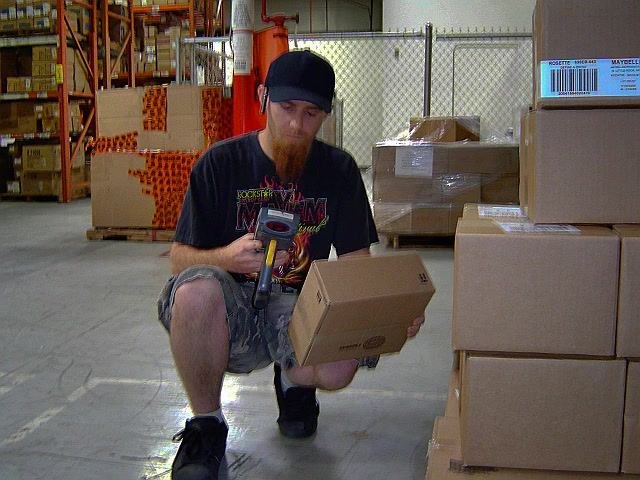 Warehouse worker using an RF scanner to scan products from an open box, surrounded by boxes and shelving