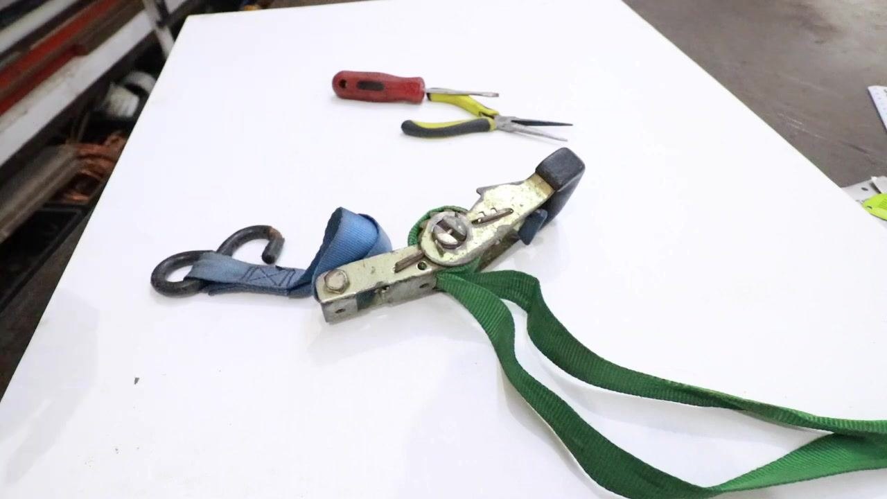 A jammed ratchet strap with green webbing placed on a white table, with needle nose pliers and a screwdriver visible in the background.