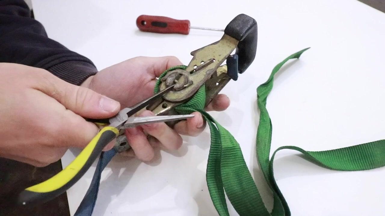Hands using needle nose pliers to rotate the center shaft of a ratchet strap, loosening green webbing, with a red-handled screwdriver visible on the table.