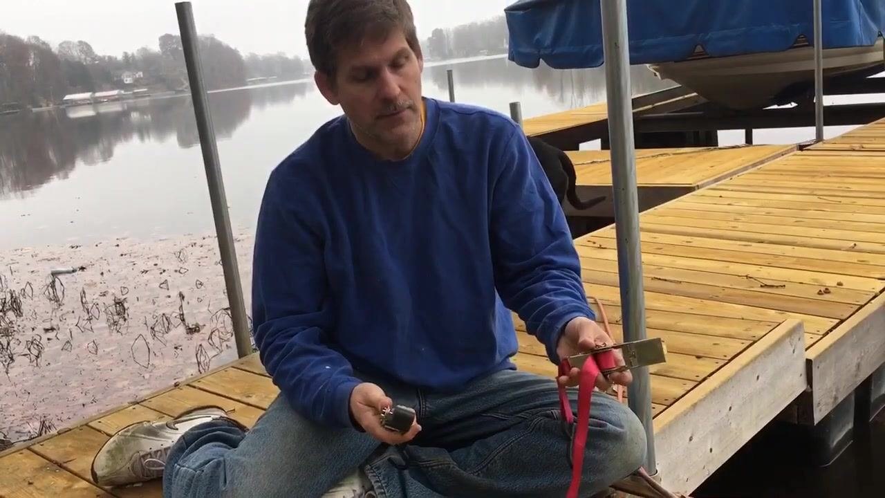 A person sits cross-legged on a wooden dock by a lake, holding a ratchet strap. Two strap types are shown, one red and one green, with a covered boat visible in the background.