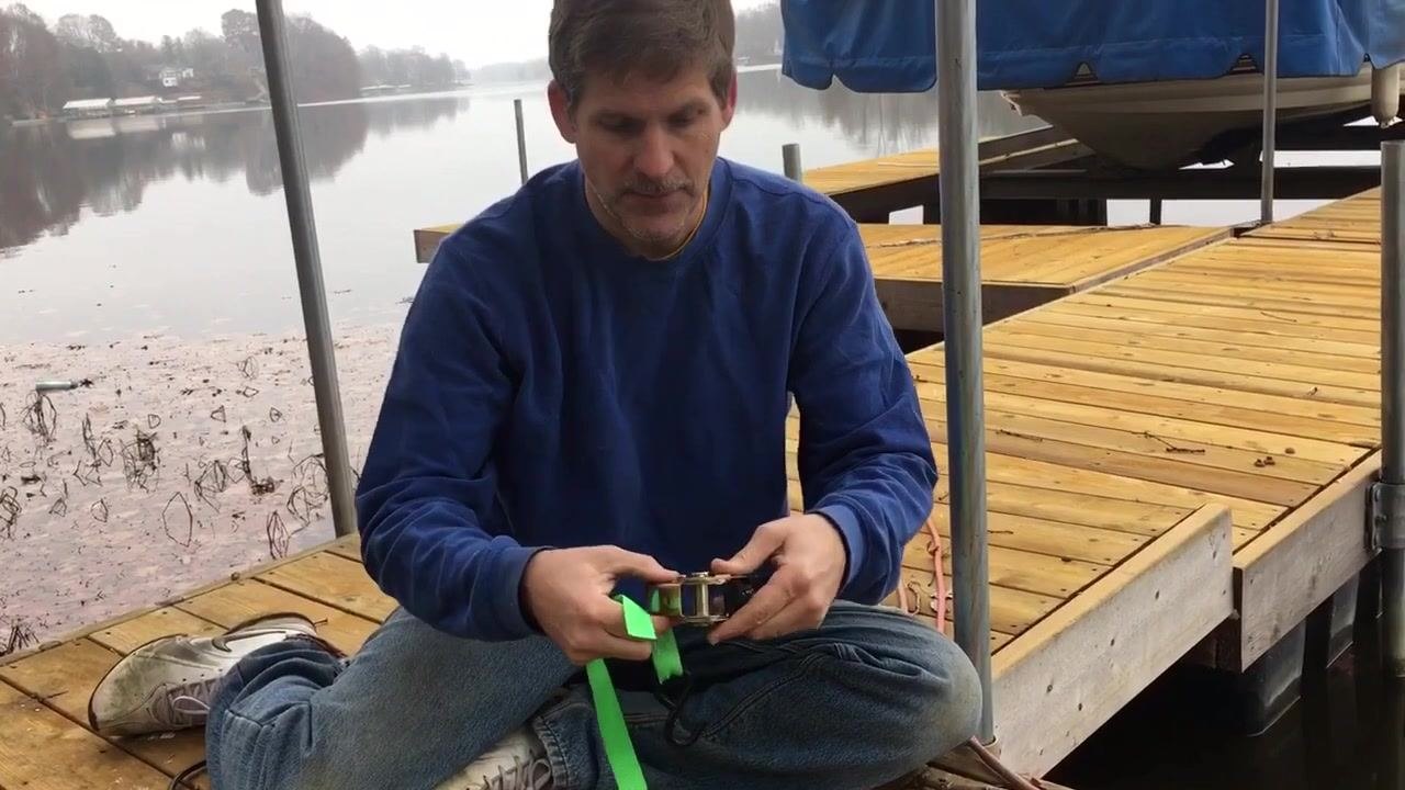 Close-up of hands threading a green strap through the barrel of a ratchet mechanism, held open on a wooden dock.
