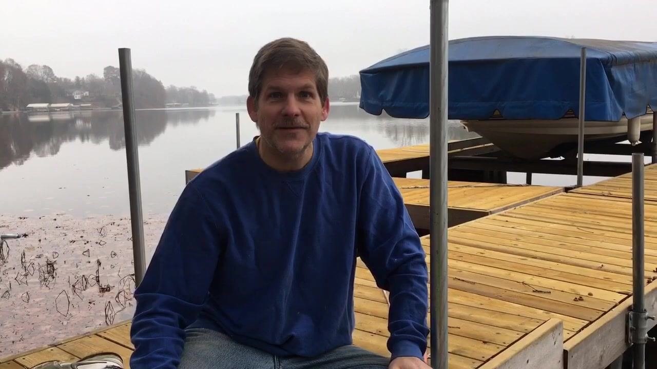 A person in a blue sweatshirt and jeans sits cross-legged on a wooden dock by a calm lake. The dock has metal posts and a covered boat is visible in the background. The person is looking at the camera, appearing to wrap up the demonstration. The environment is outdoors, overcast, and peaceful, with trees and houses visible across the lake.