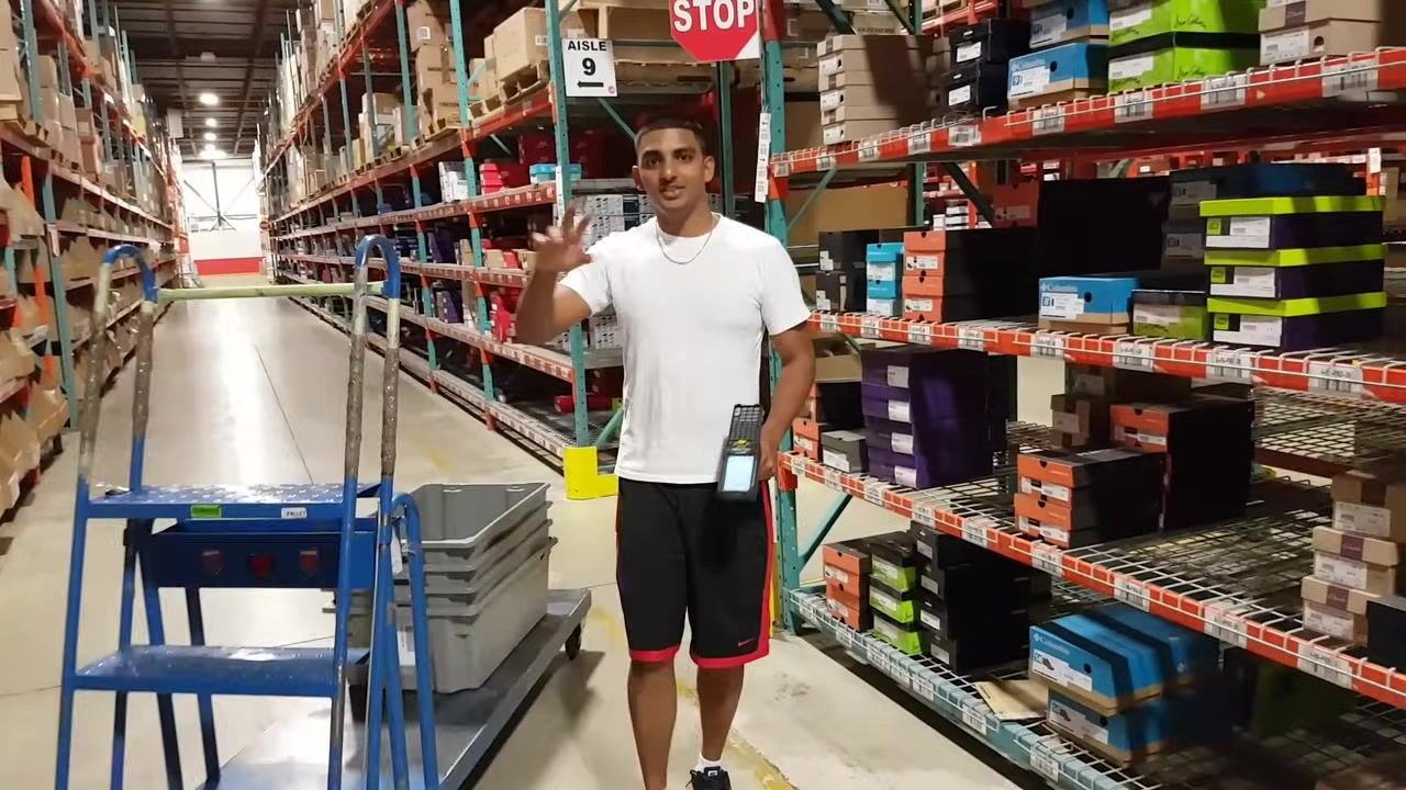 A warehouse worker stands in an aisle holding an RF gun, with shelves of shoe boxes, a blue step ladder, and a STOP sign visible.