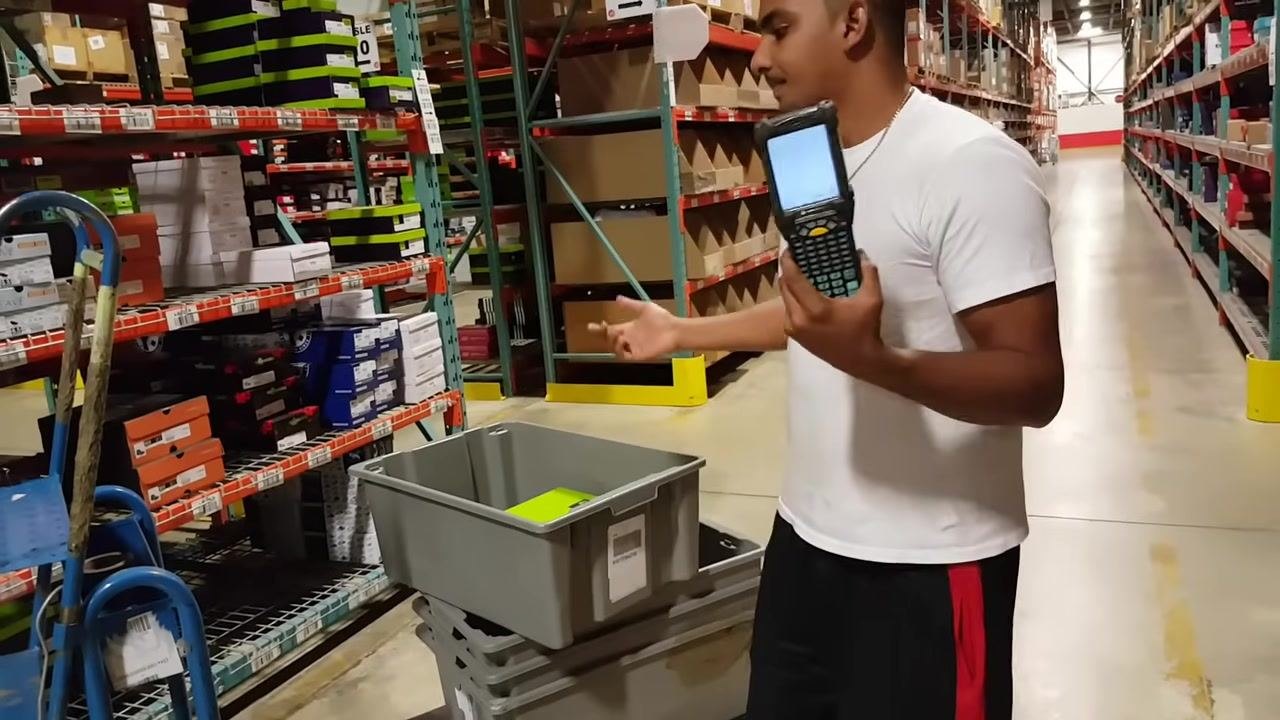 A worker in a white shirt stands in a warehouse aisle, holding a handheld scanner and gesturing towards a gray tote on a cart. Shoe boxes are visible on shelves.