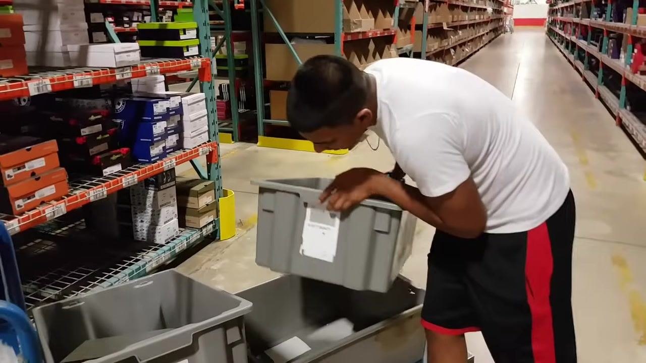 A worker in a white shirt walks back toward the warehouse shelves, holding a handheld scanner. Shoe boxes and shelf labels are visible.