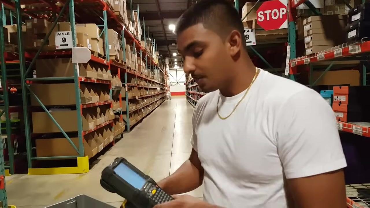 A worker in a white shirt stands in the warehouse aisle, looking at the handheld scanner. Aisle 9 signage and a stop sign are visible in the background.